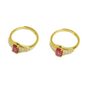 May include: Two gold-colored rings with a rectangular red gemstone and small clear stones. The rings have an adjustable band and are set against a white background. Jewelry.