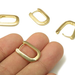 May include: Gold-colored, rectangular hoop earrings. The earrings have a simple, modern design with a smooth, polished finish. Several earrings are displayed, with one held in a hand. The earrings are a warm, metallic color.