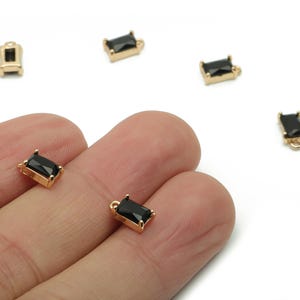 Peut inclure: Charm de bijou rectangulaire doré avec pierres noires rectangulaires. Plusieurs breloques sont éparpillées sur une surface blanche, certaines sont tenues dans une main. Les breloques ont de petites boucles pour la fixation.