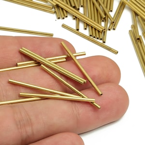 May include: A selection of small, cylindrical brass tubes. Some are held in a hand, with others scattered in the background. The tubes are a shiny gold colour and appear hollow. These components are likely used for jewellery making or other crafts.