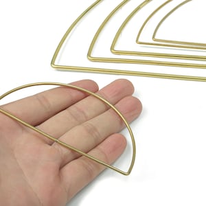 Könnte beinhalten: Eine Hand hält eine goldfarbene Halbkreisform aus Metall, mit mehreren ähnlichen Formen im Hintergrund. Die Formen bestehen aus dünnem Draht und sind halbkreisförmig angeordnet. Der Hintergrund ist weiß.