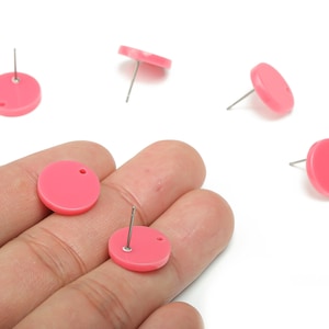 May include: Pink round stud earrings with silver posts. The earrings are a solid, vibrant pink color and have a small hole near the top. Several earrings are scattered on a white background.