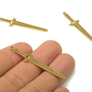 May include: Gold-coloured metal sword charms with detailed designs. The charms have a handle, a blade with etched text, and a small loop for attachment to jewellery or crafts. The image shows three charms against a white background.