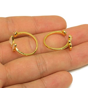 Puede incluir: Un par de pendientes de aro dorados, cada uno con un pequeño dije de mariposa rosa claro. Los pendientes se muestran sobre un fondo neutro, resaltando su diseño y color.