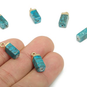 以下が含まれることがあります： ターコイズ色の六角形の宝石ペンダント。金色のキャップとループが付いています。白い背景にいくつかのペンダントが散らばっており、そのうち2つは手で持たれています。ジュエリー作りに最適です。