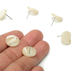 Op de afbeelding: Crèmekleurige, ronde oorstekers met een gemarmerd effect. De oorbellen hebben een pin en worden getoond in een groep van zes, waarvan sommige in een hand worden gehouden. De oorbellen zijn gemaakt van licht materiaal.
