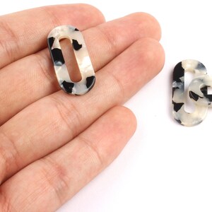 Peut inclure: Deux perles ovales à centre ouvert avec un motif écaille de tortue noir et crème. Une perle est tenue dans une main, l'autre est à côté sur une surface blanche. Ces perles sont adaptées à la fabrication de bijoux.
