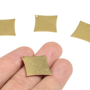 May include: Four brass-colored, square-shaped metal charms with curved sides and a small hole at the top. One charm is held in a hand. The charms have a matte finish and are likely for jewelry making or crafting.