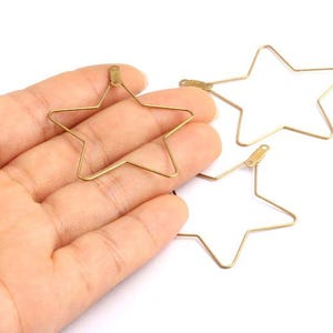 Può includere: Tre componenti per orecchini a forma di stella color oro. Uno è tenuto in mano, mentre altri due sono su una superficie bianca. Le stelle sono realizzate con filo sottile e hanno un anello nella parte superiore. Adatto per la creazione di gioielli.
