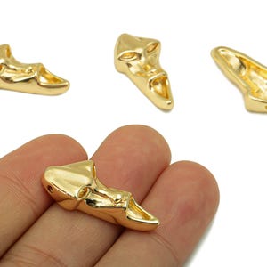 May include: Four gold-colored metal shoe-shaped charms. One charm is held in a hand, while the other three are scattered on a white background. The charms have a glossy finish and are designed with a detailed, textured surface.