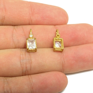 May include: Two gold-colored rectangular pendants with clear, rectangular gemstones. One pendant has a small, round gemstone above the rectangle. The pendants are displayed on a light-colored background.