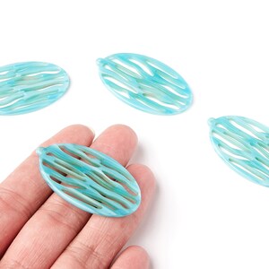Peut inclure: Quatre perles ovales turquoise avec un motif ajouré. Les perles ont un motif ondulé et un petit trou en haut. Une perle est tenue dans une main. Les perles sont sur fond blanc.