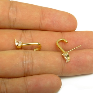 May include: A pair of gold-colored earrings, each featuring a pear-shaped, clear gemstone. The earrings have a curved design with a post for piercing. The earrings are displayed on a light-colored surface.