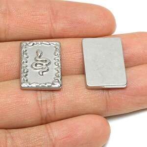 Könnte beinhalten: Zwei rechteckige Charms in Silber. Ein Charm zeigt ein erhabenes Schlangendesign in einem dekorativen Rahmen. Der andere Charm ist ein schlichtes Rechteck. Die Charms werden in einer Hand vor weißem Hintergrund gehalten.