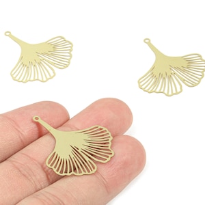 May include: Three gold-colored, ginkgo leaf-shaped charms. One charm is held in a hand, while two others are displayed on a white background. The charms have a small loop at the top for attaching to jewelry.