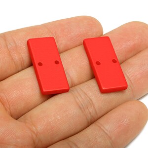 May include: Two bright red rectangular beads with two holes each. These beads are likely used for jewelry making or other craft projects. The beads are shown against a neutral background.