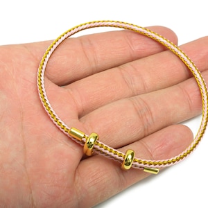 May include: A gold and pink braided bracelet with gold-colored accents. The bracelet is held in a hand, showcasing its design and craftsmanship. The bracelet's design features a woven pattern with a combination of gold and pink threads.