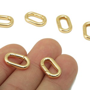 Puede incluir: Se muestran varios cierres ovalados de color dorado sobre un fondo blanco. Dos cierres se sostienen en una mano. Los cierres son de color dorado metálico brillante. Estos cierres probablemente se utilizan para la fabricación de joyas u otros proyectos de artesanía.