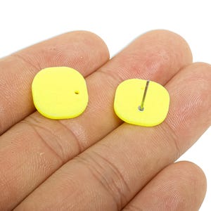May include: A pair of bright yellow square stud earrings. One earring has a small hole, and the other has a silver post for piercing. The earrings are held in a hand against a white background. Jewellery.