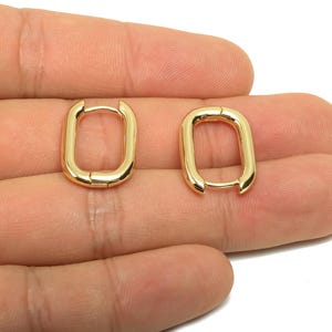 May include: A pair of gold-colored, rectangular hoop earrings. The earrings have a smooth, polished finish and a hinged closure. The earrings are displayed on a hand against a white background. Jewelry.