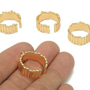 Op de afbeelding: Vier goudkleurige verstelbare ringonderdelen met een textuur van verticale balken. Eén ring wordt in een hand gehouden. Deze open ringen zijn geschikt voor het maken van doe-het-zelf sieraden.