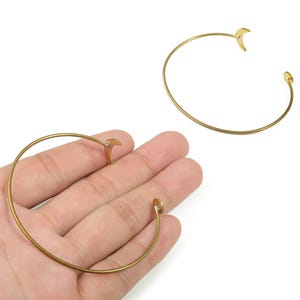 May include: Two gold-colored open cuff bracelets. One bracelet is held in a hand. Each bracelet has a curved design with a decorative element at each end. The bracelets are made of a thin, shiny metal.