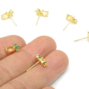May include: Gold-toned stud earrings with a cluster of green and clear gemstones. The earrings have a post backing. Several earrings are scattered on a white surface, with one held between fingers.