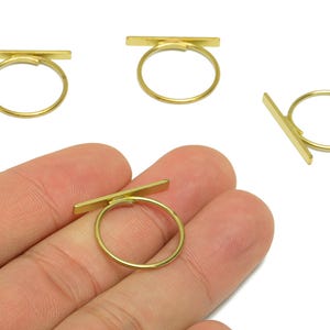 May include: Gold-colored rings with a minimalist design. Each ring features a thin, straight bar across the top of a circular band. The rings are displayed against a white background, with one ring being held by a hand.