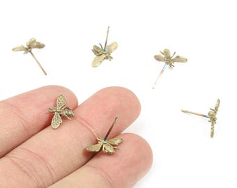 May include: A collection of small, gold-colored bee-shaped stud earrings. The earrings are detailed with wings and body segments, and are shown against a white background. The earrings are held in a hand.