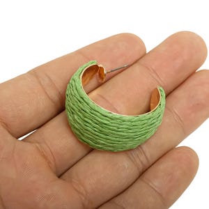 May include: A single green hoop earring with a woven texture, held in a hand. The earring has a gold-coloured interior and a silver post. The earring is a half-circle shape.