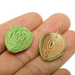 May include: A pair of green woven raffia earrings. One earring is a round shape, and the other is a gold-coloured metal teardrop shape with a post for piercing. The earrings are displayed on a hand.