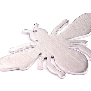 Peut inclure: Une découpe de métal argentée en forme d'abeille avec une finition brossée. L'abeille est en position de vol, les ailes déployées.