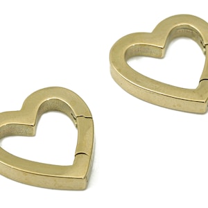 Könnte beinhalten: Zwei goldfarbene herzförmige Metallverschlüsse. Die Verschlüsse sind offen und bereit, an einer Tasche oder einem anderen Gegenstand befestigt zu werden.
