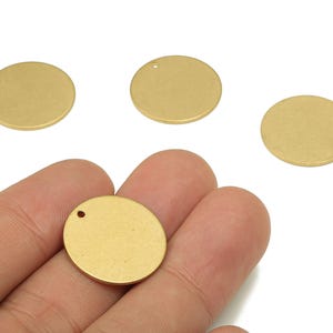 以下が含まれることがあります： 小さな穴の開いた、4つの金色の丸い金属製のチャーム。1つのチャームが手に持たれています。チャームは明るくマットな金色で、白い背景にあります。これらのチャームは、ジュエリー作りに最適です。