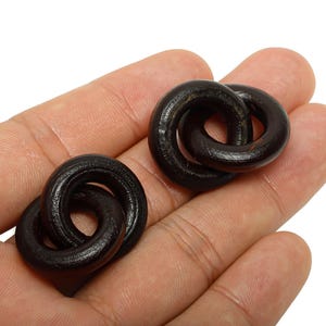 Könnte beinhalten: Zwei dunkelbraune Holzringe, die ineinandergreifen, jeweils mit kreisförmiger Form, werden in der Hand einer Person gehalten. Die Ringe sind ineinander verschlungen und bilden ein kettenartiges Design. Das Holz hat ein glattes, poliertes Aussehen.