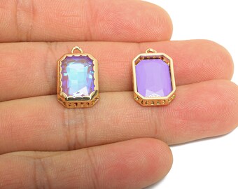 May include: Two rectangular gold-colored pendants with a loop at the top. One pendant has an iridescent, multi-colored gem, and the other has a solid lavender gem. The gems are set in a gold-colored frame with a decorative edge.