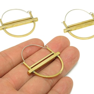 以下が含まれることがあります： ハーフサークルとストレートバーのデザインが特徴の幾何学的な真鍮製イヤリング。イヤリングはゴールド仕上げで、シルバーカラーのフックが付いています。画像には3つのイヤリングが写っており、そのうちの1つは手に持たれています。