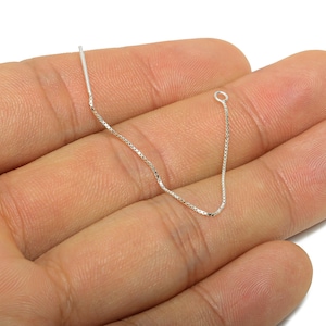 Può includere: Un delicato orecchino a catena d'argento con un piccolo anello a un'estremità e un perno dritto all'altra. La catena è tenuta in mano, mostrando il suo design a maglie quadrate. L'orecchino è probabilmente per orecchie forate.