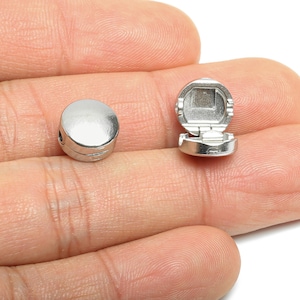 May include: Two small, silver-coloured metal jewellery findings. One is a round, hinged clasp, open to reveal a square interior. The other is a matching, closed round finding. Both are shown against a neutral background.