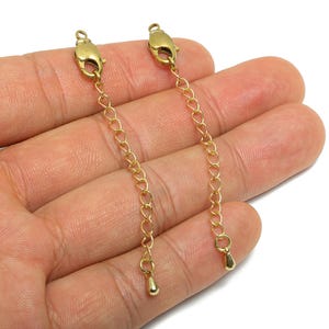 May include: Two gold-coloured chain extenders with lobster claw clasps and teardrop charms. The extenders are made of small, heart-shaped links. The clasps are attached to the top, and the charms hang from the bottom.