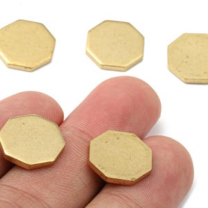 May include: Close-up of several small, gold-colored, octagonal metal charms. Two charms are held between fingers, while three others are scattered nearby. The charms have a matte finish and are likely used for jewelry making or crafting.