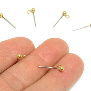 May include: Close-up of several gold-coloured ball head pins with silver-coloured stems. The pins have a small loop at the end of the gold ball. The pins are on a white background.