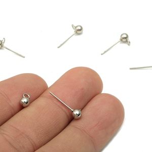 Pode incluir: Achados de joalheria prateados, incluindo vários pinos com ponta de bola e um pequeno berloque com uma argola. Os achados são exibidos em um fundo branco, alguns sendo segurados em uma mão.