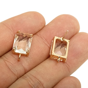May include: Two gold-toned rectangular earrings with clear, faceted, rectangular stones. One earring has a post, the other has a loop. The earrings are held in a hand against a white background. Jewelry.