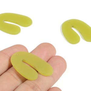 Puede incluir: Tres colgantes acrílicos en forma de U de color verde claro. Uno de los colgantes se sostiene en una mano, mientras que otros dos están dispersos sobre un fondo blanco. Cada colgante tiene un pequeño agujero en la parte superior para ensartar.