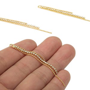 May include: Gold-colored jewelry findings with three strands of small, clear crystals. The findings have a loop at the top for attachment and a beaded chain at the bottom. The jewelry is shown against a white background.