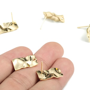 May include: Gold-colored stud earrings with a textured, wavy design. The earrings have a post backing and a shiny finish. The image shows several pairs of the earrings, with some held in a hand.