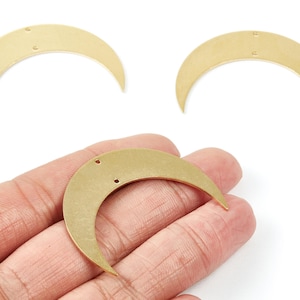 Puede incluir: Tres dijes de luna creciente de color latón. Dos se muestran por separado y uno se sostiene en una mano. Cada dije tiene dos pequeños agujeros para ensartar. Los dijes tienen un acabado liso y mate, ideal para la fabricación de joyas.