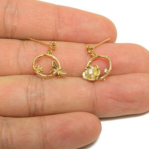 May include: A pair of gold-colored earrings. One earring features a small flower and a butterfly within a circular frame. The other earring has a flower with a central gem and a small star-shaped accent. Both earrings have a post for pierced ears.