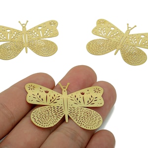 May include: Gold-colored butterfly-shaped metal charms with intricate cut-out designs. The butterfly wings feature floral and circular patterns. One charm is held in a hand, with two others visible in the background. These charms are suitable for jewelry making.